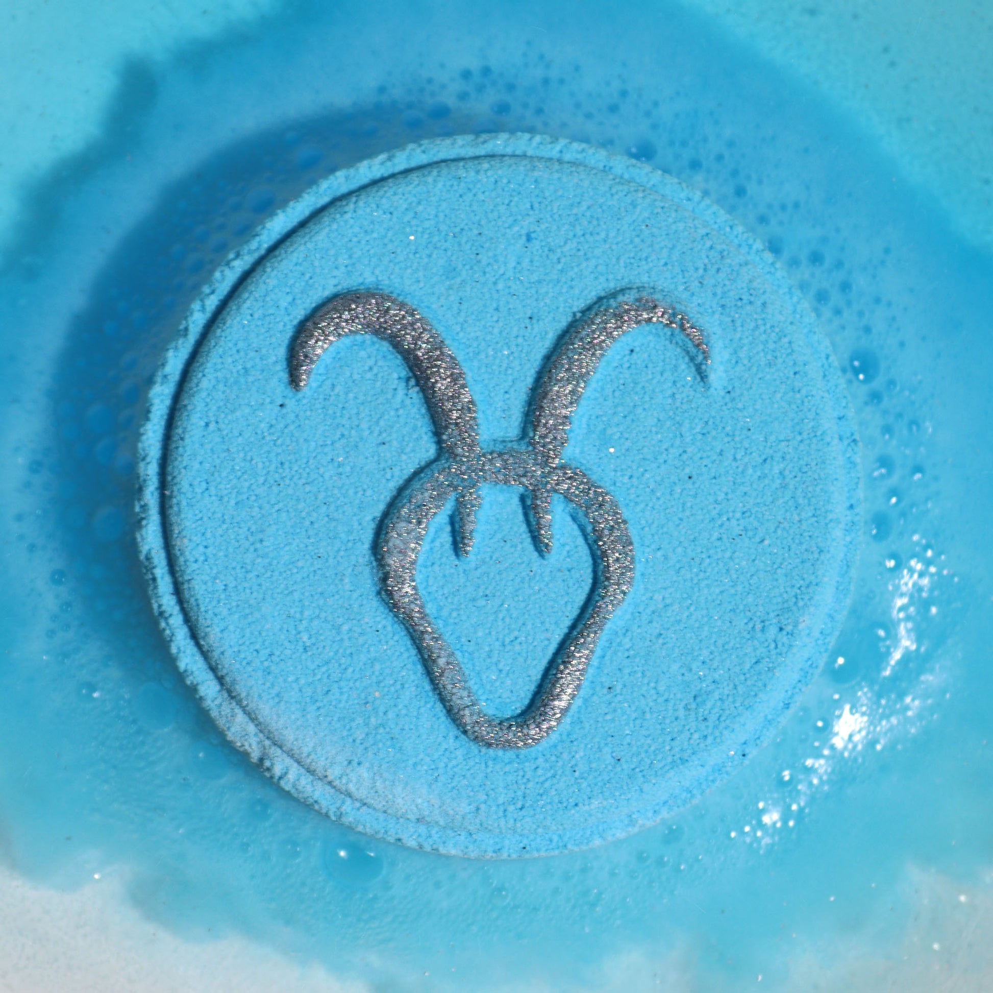 Close-up of Capricorn bath bomb fizzing with grounding fragrance, a silver symbol on a circular light blue bath bomb