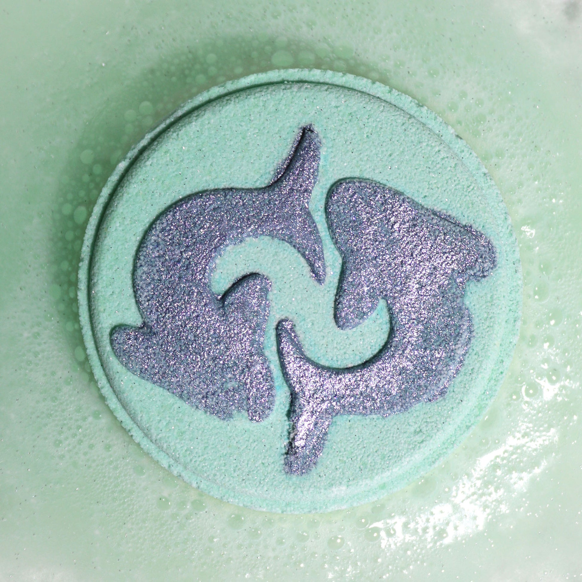 Close-up of Pisces bath bomb fizzing with soft, dreamy colours and gentle floral fragrance, with a dolphin design on a light green circular bath bomb