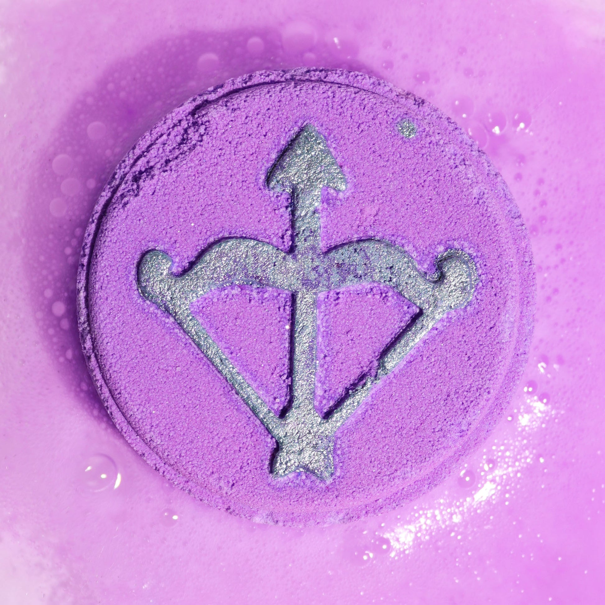 Close-up of Sagittarius silver symbol on a purple circular bath bomb fizzing in bright colours with uplifting citrus fragrance.