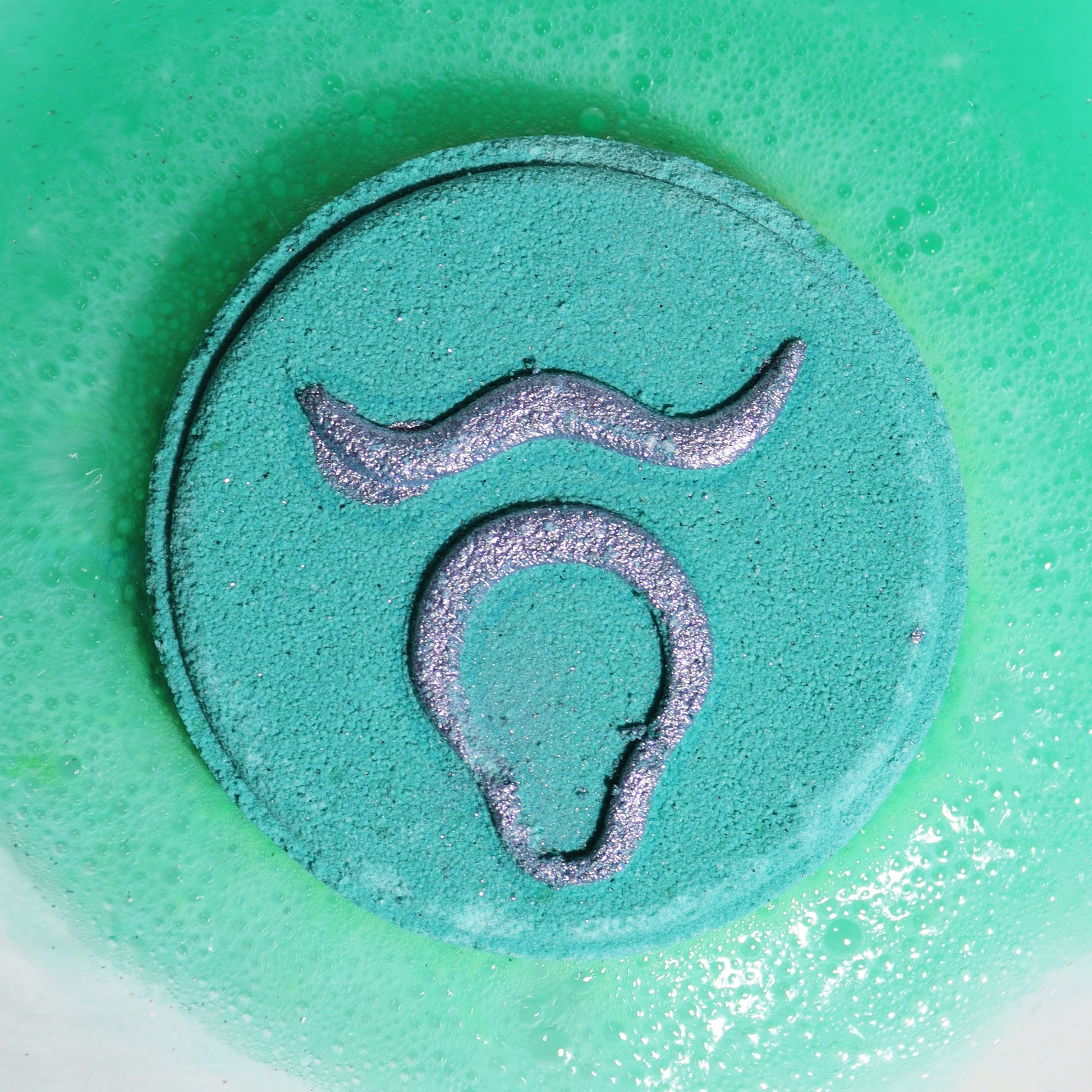 Close-up of Taurus bath bomb fizzing in bathwater with rich green colours and grounding fragrance notes of patchouli and amber.
